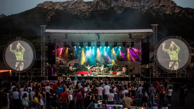 Festival de Musique Voix de Femmes à Maury en plein air au pied des montagnes, avec un groupe de musiciens sur scène et un public rassemblé devant. Deux grands écrans circulaires de chaque côté projettent l’image de la chanteuse Cléa Vincent en train de se produire.