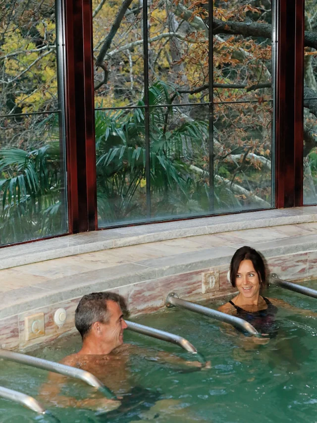 Deux personnes se détendent dans un bassin thermal intérieur avec jets d’eau, face à une grande baie vitrée donnant sur un paysage arboré.