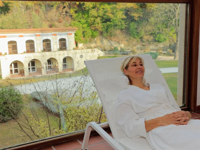 Une femme en peignoir se détend sur un transat à l’intérieur des Thermes du Boulou, profitant d’une vue apaisante sur le parc thermal et le bâtiment principal à travers une large baie vitrée.