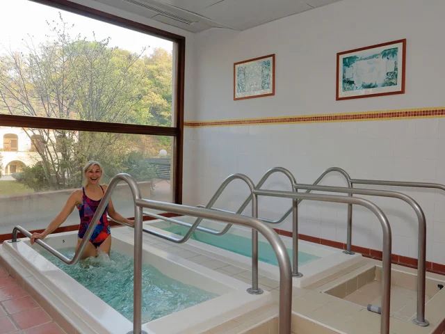 Une femme souriante effectue une séance de marche en bassin thermal, tenant les rampes métalliques, dans une salle lumineuse donnant sur un jardin arboré.