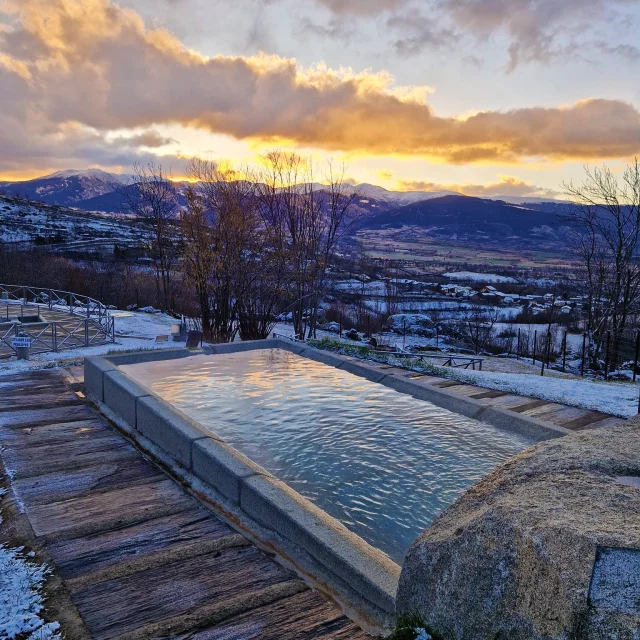 Bassin extérieur des Bains de Dorres en hiver, entouré de neige, avec vue sur les montagnes enneigées et un coucher de soleil doré derrière les nuages.