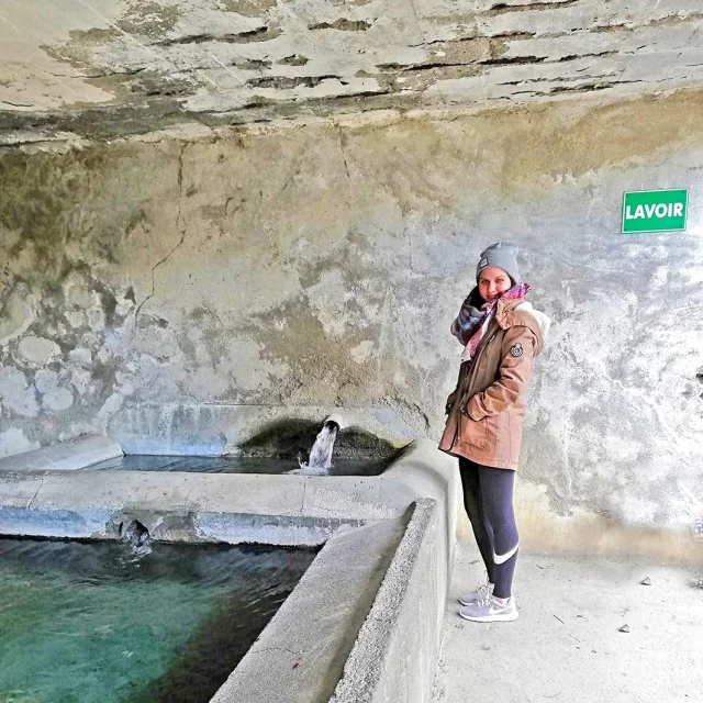 Une femme souriante se tient près du lavoir des Bains de Dorres, à côté d’un bassin en pierre alimenté par une source d’eau thermale. Elle porte un manteau marron, un bonnet gris et une écharpe colorée.
