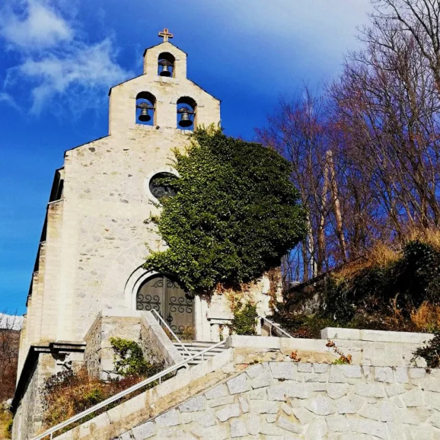 Façade de l’église romane Saint-Fructueux à Llo, en pierre claire, ornée de trois cloches sous un ciel bleu, avec du lierre grimpant sur le mur.