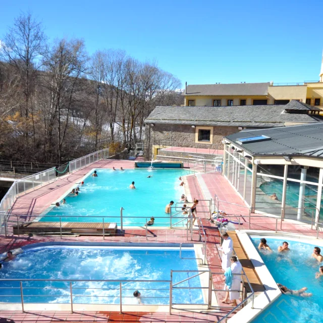 Vue des bassins extérieurs des Bains de Llo par temps ensoleillé, avec plusieurs personnes profitant des eaux thermales entourées de montagnes et d’arbres.