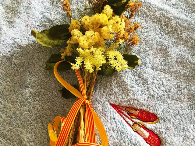 Bouquet traditionnel de la Sant Joan composé de fleurs jaunes et de plantes séchées, noué d’un ruban catalan rouge et or, posé sur un tissu bleu brodé d’espadrilles rouges catalanes.