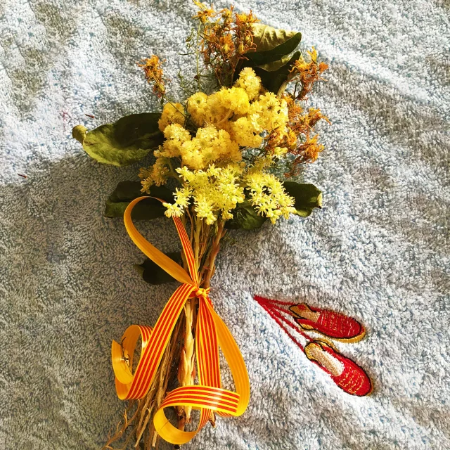 Bouquet traditionnel de la Sant Joan composé de fleurs jaunes et de plantes séchées, noué d’un ruban catalan rouge et or, posé sur un tissu bleu brodé d’espadrilles rouges catalanes.