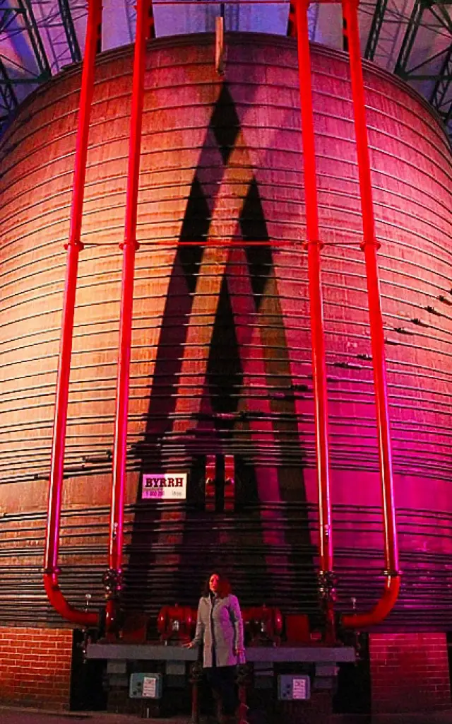 Vue impressionnante de la plus grande cuve en chêne du monde à la Cave Byrrh de Thuir, illuminée de couleurs vives, avec une personne au premier plan soulignant l’échelle monumentale du lieu.