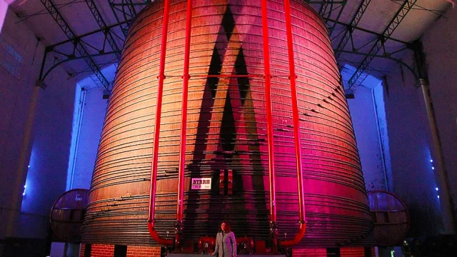 Vue impressionnante de la plus grande cuve en chêne du monde à la Cave Byrrh de Thuir, illuminée de couleurs vives, avec une personne au premier plan soulignant l’échelle monumentale du lieu.