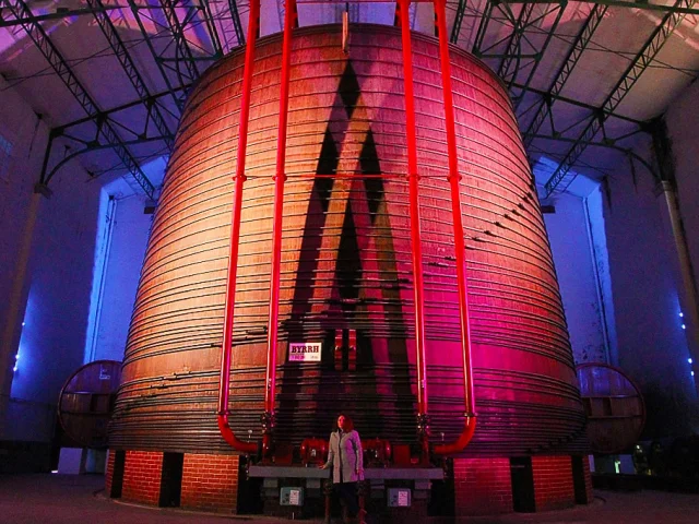 Vue impressionnante de la plus grande cuve en chêne du monde à la Cave Byrrh de Thuir, illuminée de couleurs vives, avec une personne au premier plan soulignant l’échelle monumentale du lieu.