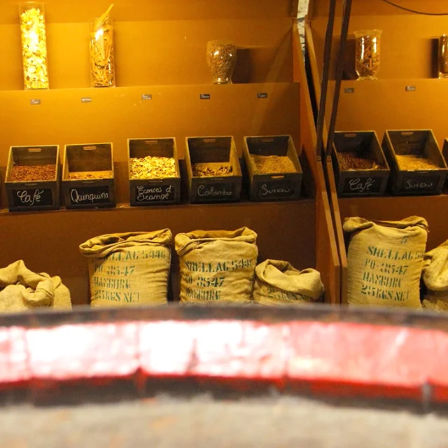 Présentation des ingrédients utilisés dans la fabrication du Byrrh à la Cave Byrrh de Thuir : bocaux et caisses remplis d’épices, d’écorces d’orange, de cannelle, de cacao, de café et de quinquina, disposés sur un présentoir accompagné de sacs en toile.