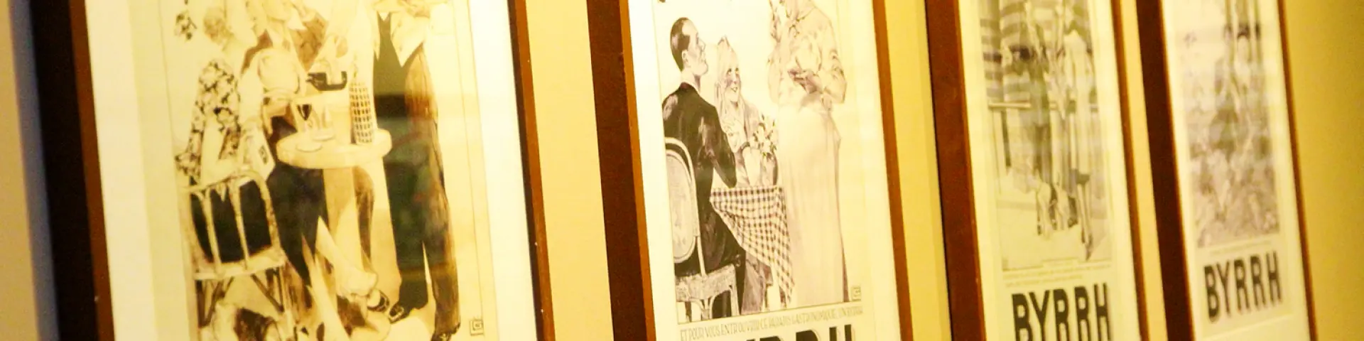 Série d’affiches publicitaires anciennes en noir et blanc de la marque Byrrh, encadrées et exposées sur un mur à la Cave Byrrh de Thuir, illustrant l’évolution du style graphique et de la communication de la marque au fil du temps.