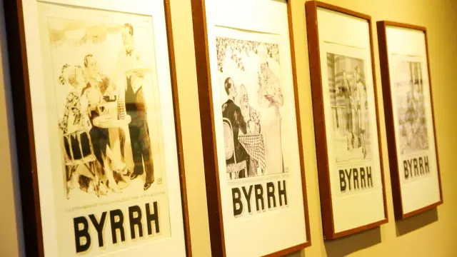 Série d’affiches publicitaires anciennes en noir et blanc de la marque Byrrh, encadrées et exposées sur un mur à la Cave Byrrh de Thuir, illustrant l’évolution du style graphique et de la communication de la marque au fil du temps.