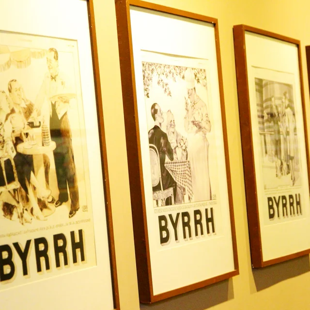 Série d’affiches publicitaires anciennes en noir et blanc de la marque Byrrh, encadrées et exposées sur un mur à la Cave Byrrh de Thuir, illustrant l’évolution du style graphique et de la communication de la marque au fil du temps.