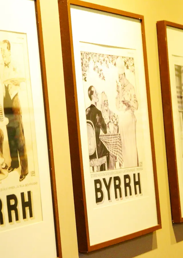 Série d’affiches publicitaires anciennes en noir et blanc de la marque Byrrh, encadrées et exposées sur un mur à la Cave Byrrh de Thuir, illustrant l’évolution du style graphique et de la communication de la marque au fil du temps.