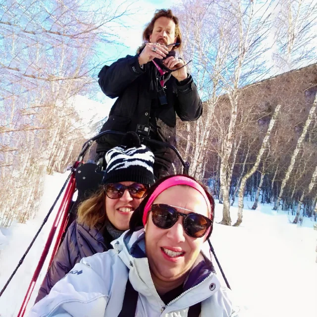 Deux femmes souriantes assises dans un traîneau à chiens en montagne, avec leur guide debout derrière elles, entourés de neige et d’arbres nus.