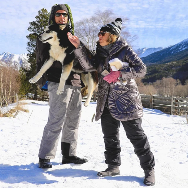 Moment de complicité entre un musher, une femme et un husky dans un paysage enneigé de montagne.