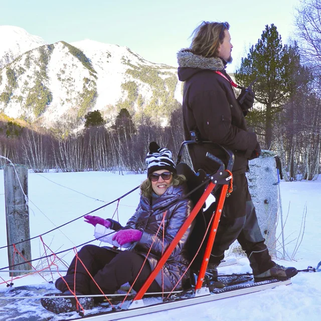 Balade en chiens de traîneau : une femme souriante assise sur le traîneau, accompagnée du musher, dans un paysage enneigé de montagne.