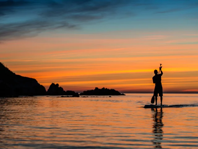 Silhouette d’une personne pratiquant le paddle sur une mer calme au coucher du soleil, avec un ciel orangé et bleu se reflétant sur l’eau.