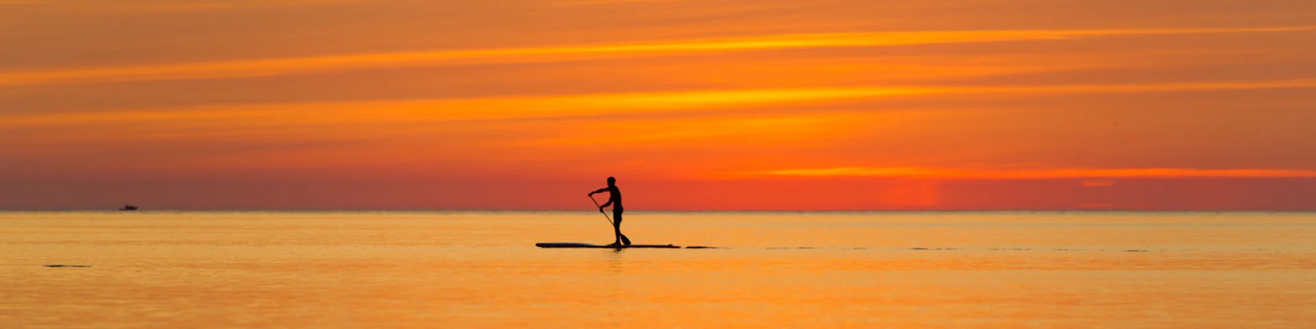 Une personne en silhouette pratique le paddle sur une mer calme au coucher du soleil, sous un ciel aux teintes orange et dorées.