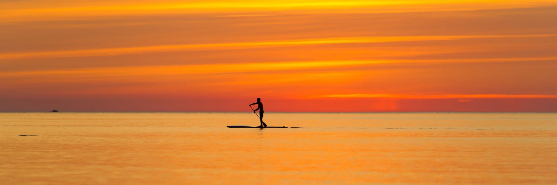 Une personne en silhouette pratique le paddle sur une mer calme au coucher du soleil, sous un ciel aux teintes orange et dorées.
