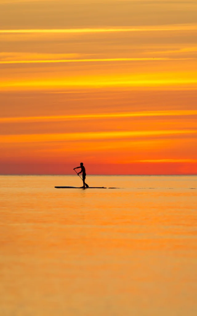 Une personne en silhouette pratique le paddle sur une mer calme au coucher du soleil, sous un ciel aux teintes orange et dorées.