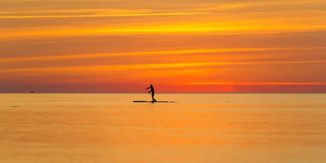 Une personne en silhouette pratique le paddle sur une mer calme au coucher du soleil, sous un ciel aux teintes orange et dorées.