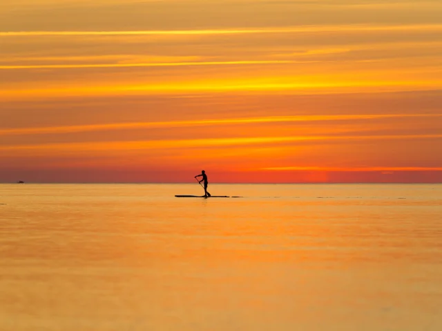 Une personne en silhouette pratique le paddle sur une mer calme au coucher du soleil, sous un ciel aux teintes orange et dorées.