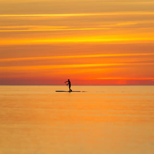 Une personne en silhouette pratique le paddle sur une mer calme au coucher du soleil, sous un ciel aux teintes orange et dorées.