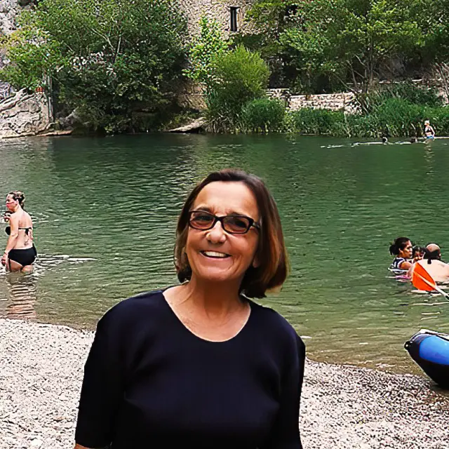 Femme souriante au bord d’une rivière fréquentée par des baigneurs et des enfants en canoë.