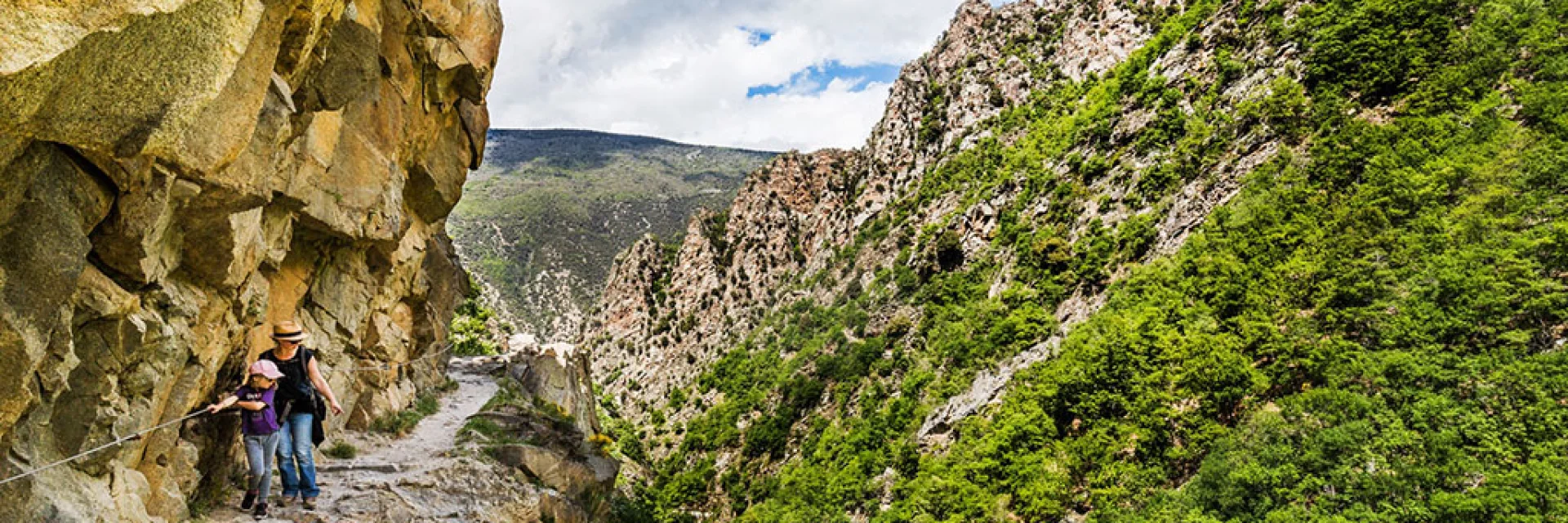 Deux randonneuses avancent prudemment sur un sentier étroit taillé dans la falaise, longeant une paroi rocheuse vertigineuse des Gorges de la Carança, entourée de montagnes verdoyantes sous un ciel partiellement nuageux.