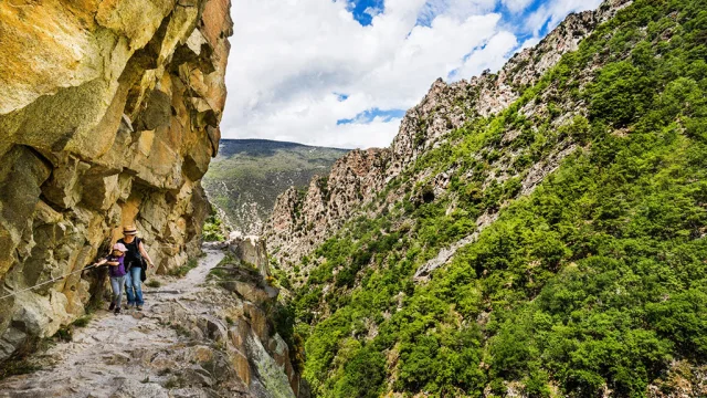 Deux randonneuses avancent prudemment sur un sentier étroit taillé dans la falaise, longeant une paroi rocheuse vertigineuse des Gorges de la Carança, entourée de montagnes verdoyantes sous un ciel partiellement nuageux.