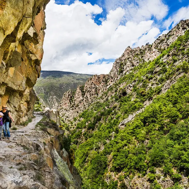 Deux randonneuses avancent prudemment sur un sentier étroit taillé dans la falaise, longeant une paroi rocheuse vertigineuse des Gorges de la Carança, entourée de montagnes verdoyantes sous un ciel partiellement nuageux.