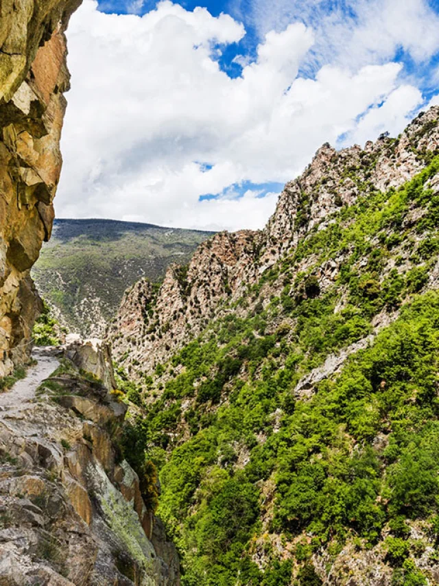 Deux randonneuses avancent prudemment sur un sentier étroit taillé dans la falaise, longeant une paroi rocheuse vertigineuse des Gorges de la Carança, entourée de montagnes verdoyantes sous un ciel partiellement nuageux.