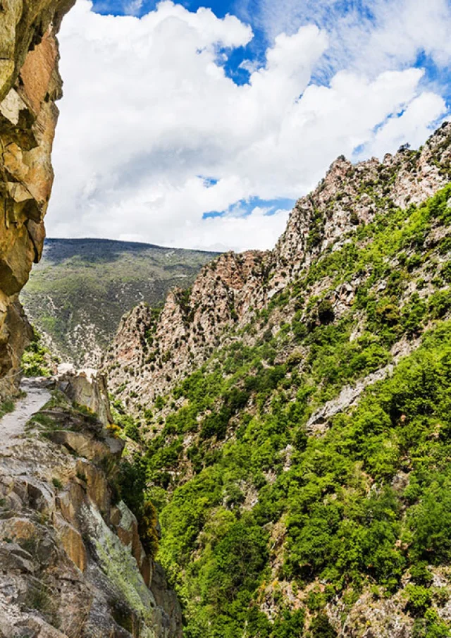 Deux randonneuses avancent prudemment sur un sentier étroit taillé dans la falaise, longeant une paroi rocheuse vertigineuse des Gorges de la Carança, entourée de montagnes verdoyantes sous un ciel partiellement nuageux.