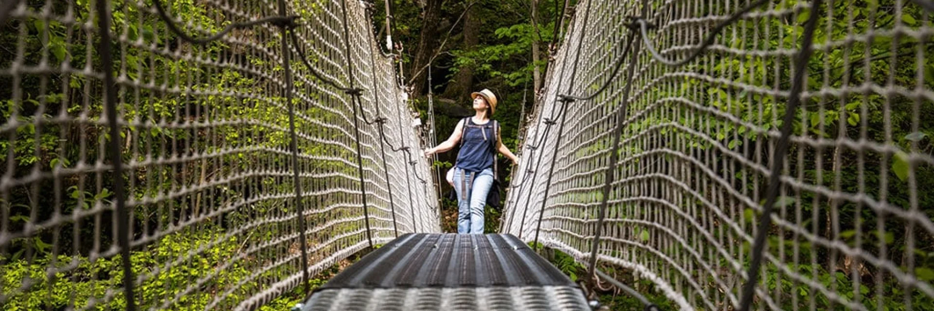 Femme marchant sur un pont suspendu métallique entouré de filets de sécurité, au-dessus d’une gorge boisée des Gorges de la Carança, sous une lumière naturelle filtrée par la végétation.