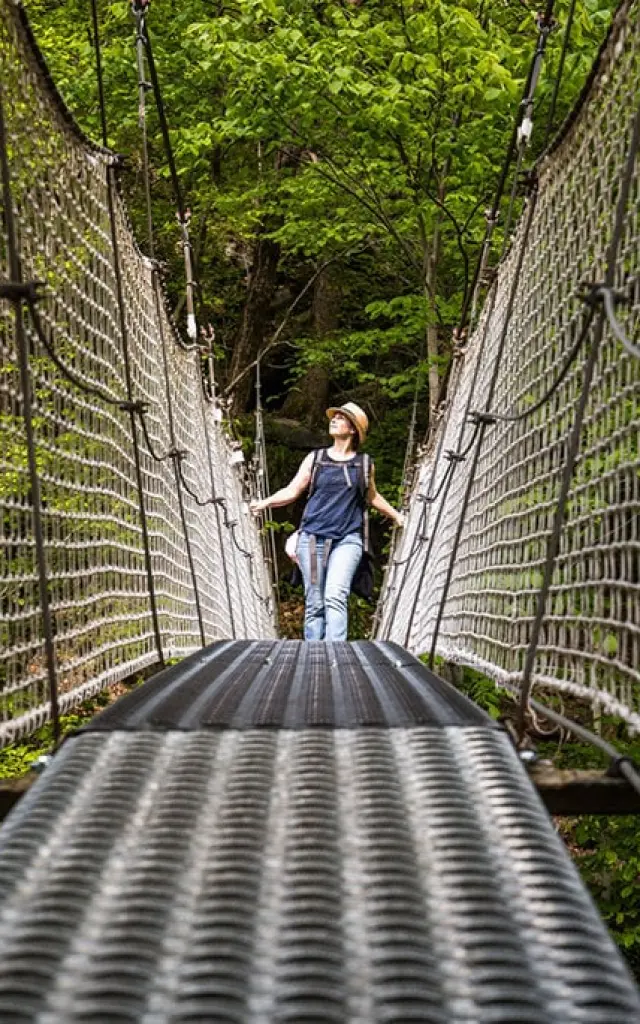 Femme marchant sur un pont suspendu métallique entouré de filets de sécurité, au-dessus d’une gorge boisée des Gorges de la Carança, sous une lumière naturelle filtrée par la végétation.