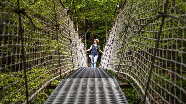 Femme marchant sur un pont suspendu métallique entouré de filets de sécurité, au-dessus d’une gorge boisée des Gorges de la Carança, sous une lumière naturelle filtrée par la végétation.