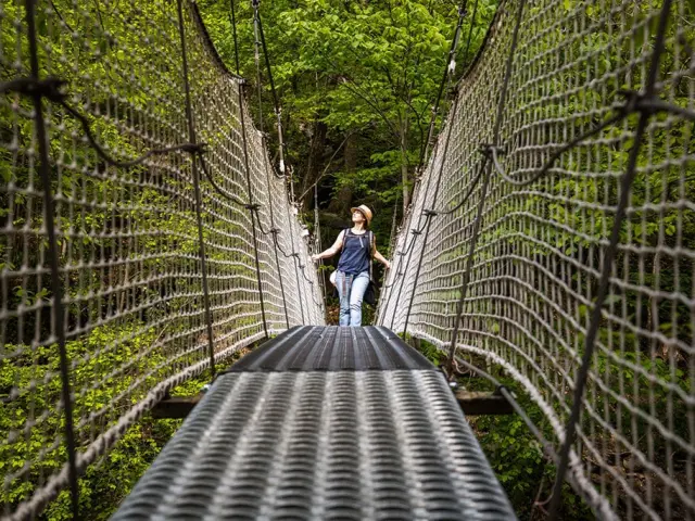 Femme marchant sur un pont suspendu métallique entouré de filets de sécurité, au-dessus d’une gorge boisée des Gorges de la Carança, sous une lumière naturelle filtrée par la végétation.