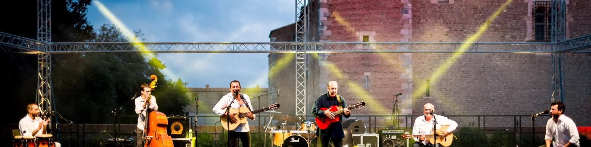 Un groupe de six musiciens joue sur scène en plein air devant le Palais des Rois de Majorque à Perpignan, sous un éclairage de scène jaune et blanc. Plusieurs instruments sont visibles, dont des guitares, des percussions, une contrebasse et un cajón.