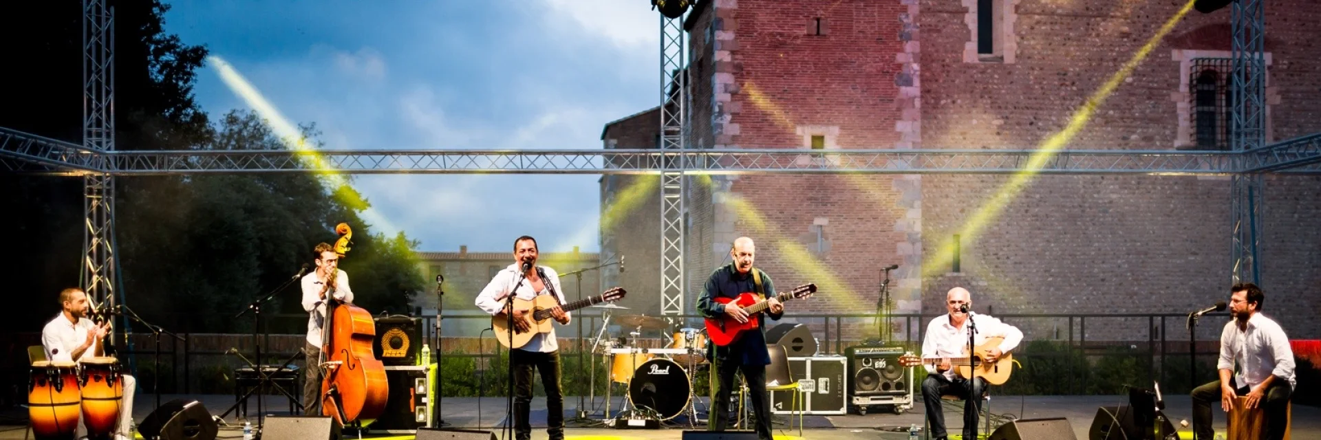 Un groupe de six musiciens joue sur scène en plein air devant le Palais des Rois de Majorque à Perpignan, sous un éclairage de scène jaune et blanc. Plusieurs instruments sont visibles, dont des guitares, des percussions, une contrebasse et un cajón.