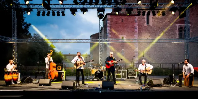 Un groupe de six musiciens joue sur scène en plein air devant le Palais des Rois de Majorque à Perpignan, sous un éclairage de scène jaune et blanc. Plusieurs instruments sont visibles, dont des guitares, des percussions, une contrebasse et un cajón.