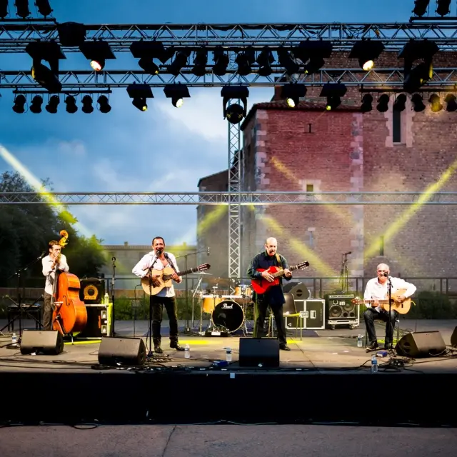 Un groupe de six musiciens joue sur scène en plein air devant le Palais des Rois de Majorque à Perpignan, sous un éclairage de scène jaune et blanc. Plusieurs instruments sont visibles, dont des guitares, des percussions, une contrebasse et un cajón.