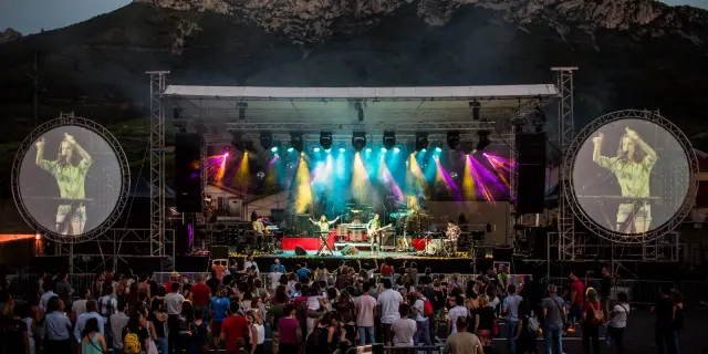 Festival de Musique Voix de Femmes à Maury en plein air au pied des montagnes, avec un groupe de musiciens sur scène et un public rassemblé devant. Deux grands écrans circulaires de chaque côté projettent l’image de la chanteuse Cléa Vincent en train de se produire.