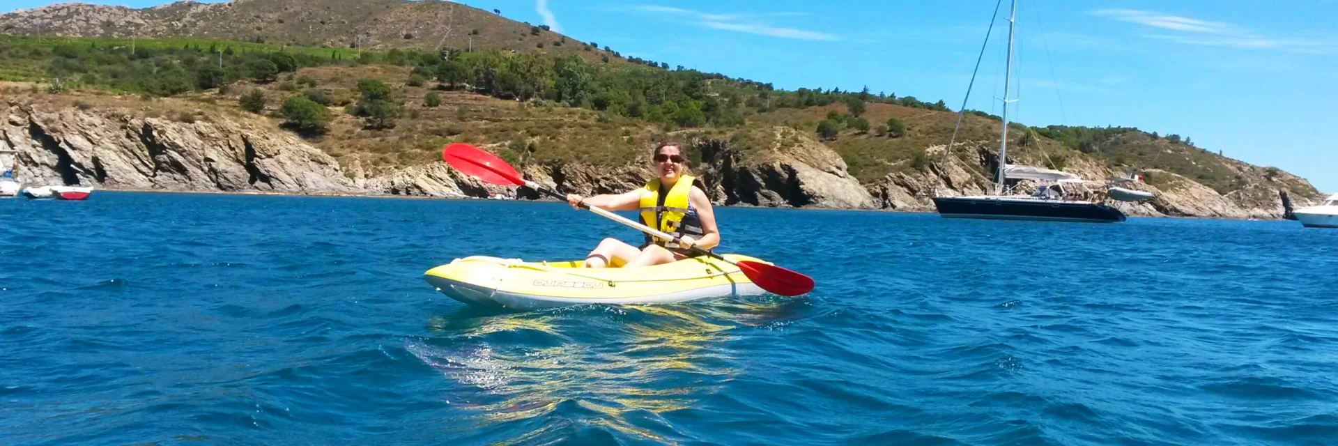 Femme pagayant en kayak jaune sur la mer à Paulilles, sous un ciel bleu, avec la côte rocheuse et des voiliers au large en arrière-plan.