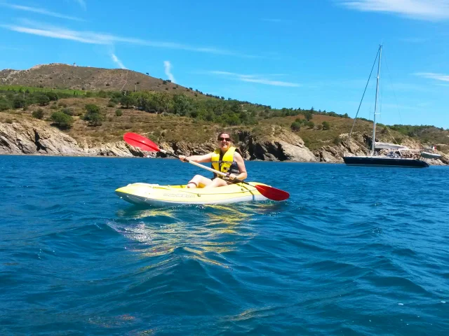 Femme pagayant en kayak jaune sur la mer à Paulilles, sous un ciel bleu, avec la côte rocheuse et des voiliers au large en arrière-plan.