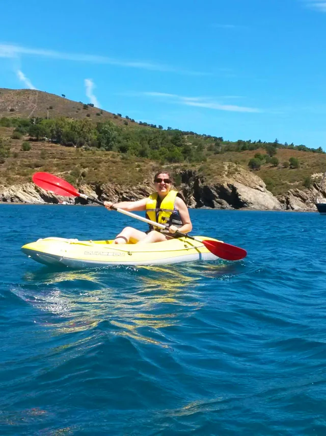 Femme pagayant en kayak jaune sur la mer à Paulilles, sous un ciel bleu, avec la côte rocheuse et des voiliers au large en arrière-plan.