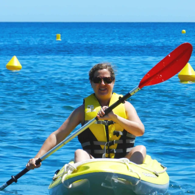 Femme souriante pagayant en kayak jaune sur la mer à Paulilles, portant un gilet de sauvetage jaune sous un ciel bleu.