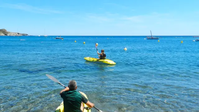 Deux personnes partent en kayak jaune depuis la plage de Paulilles, pagayant sur une mer calme et bleue sous un ciel ensoleillé.