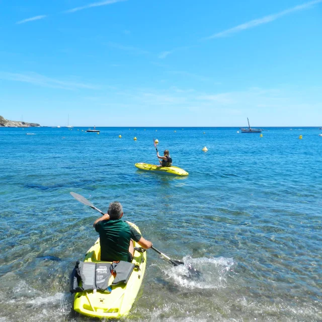 Deux personnes partent en kayak jaune depuis la plage de Paulilles, pagayant sur une mer calme et bleue sous un ciel ensoleillé.