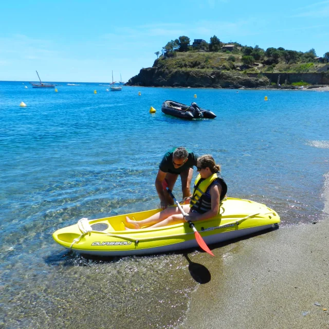 Un homme donne des explications à une femme assise dans un kayak jaune au bord de la mer, sur la plage de Paulilles, par temps ensoleillé.